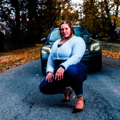 A woman in a light sweater and heels poses confidently while crouching on a street covered with autumn leaves, with a car parked behind her. The scene captures a vibrant fall atmosphere.
