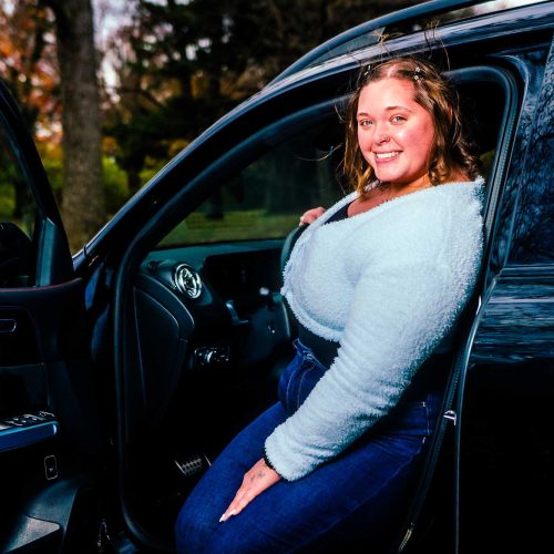 A young woman is smiling while sitting on the edge of a car door, with a scenic background of trees. The photo captures a cheerful moment outdoors.