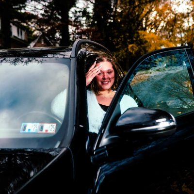 A woman smiles while sitting in a black car with the door open, surrounded by autumn foliage. The scene conveys a sense of warmth and casual comfort.