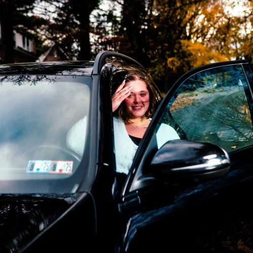 A woman smiles while sitting in a black car with the door open, surrounded by autumn foliage. The scene conveys a sense of warmth and casual comfort.