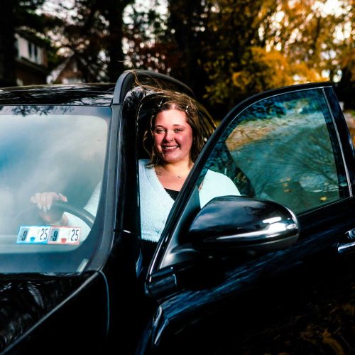 A young woman smiles while sitting in the driver's seat of a black car parked on a tree-lined street with autumn leaves. The scene captures a cheerful moment in a vibrant fall setting.