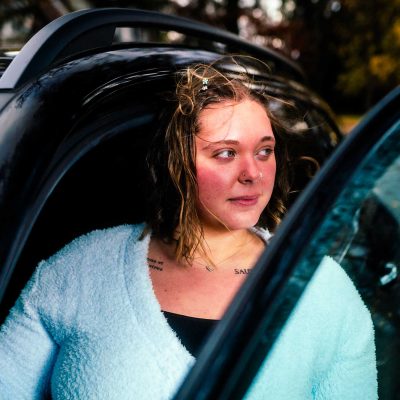 A woman with windblown hair in a fluffy blue sweater stands by an open car door, gazing thoughtfully into the distance. The scene is set outdoors.