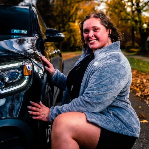 A young woman smiles while kneeling next to a black car, surrounded by autumn foliage. The scene captures a cheerful moment outdoors.