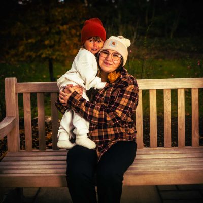 A woman sits on a wooden bench holding a young child, both wearing warm hats, in a natural outdoor setting. The scene conveys a sense of warmth and affection.