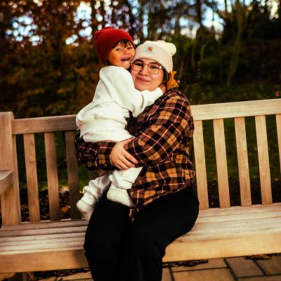 A woman sits on a bench in a park, holding a child who is smiling and wearing a red hat. The scene is set against a backdrop of autumn foliage.