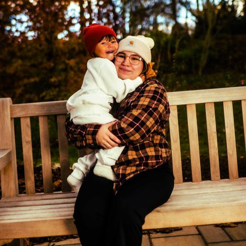 A woman sits on a bench in a park, holding a child who is smiling and wearing a red hat. The scene is set against a backdrop of autumn foliage.
