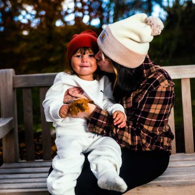 A mother kisses her child while sitting on a bench in a park, surrounded by autumn foliage. The child is wearing a white outfit and a red hat, while the mother is dressed in a plaid shirt and a knitted beanie.