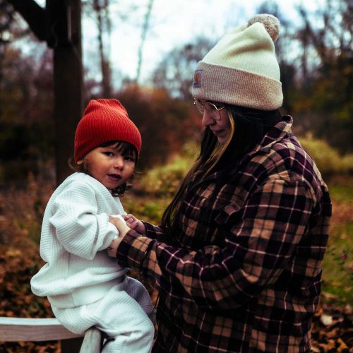 A child wearing a red beanie sits on a bench while being held by an adult in a plaid jacket, surrounded by autumn foliage. The scene captures a warm moment in a natural setting.