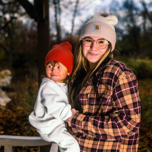 A young woman holds a smiling child in a cozy outdoor setting, both wearing warm hats. The background features trees and autumn foliage.