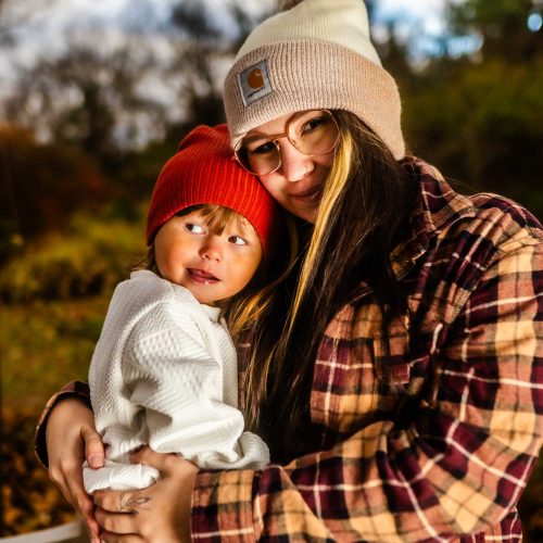 A woman with long hair and glasses smiles while holding a young child wearing a red beanie. They are surrounded by a natural setting with autumn colors.