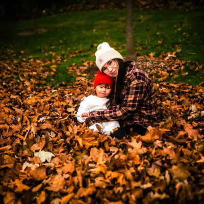A woman and a child sit together in a pile of autumn leaves, both wearing warm hats. The scene captures a cozy moment in a colorful fall setting.