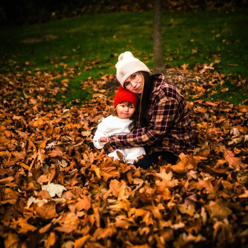 A woman and a child sit together in a pile of autumn leaves, both wearing warm hats. The scene captures a cozy moment in a colorful fall setting.