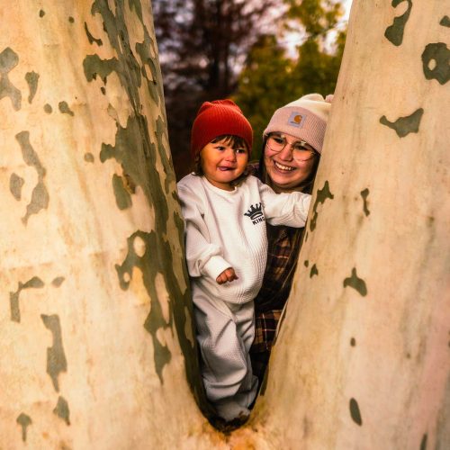 A woman and a child are playfully peeking out from between the trunks of two trees, both smiling and wearing warm hats. The scene captures a joyful moment in a natural setting.