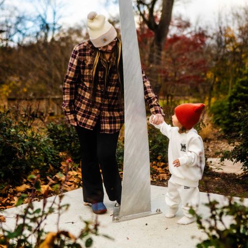 A woman and a child are standing in a garden, interacting with a tall, white monument. The scene is set in autumn, with colorful foliage in the background.